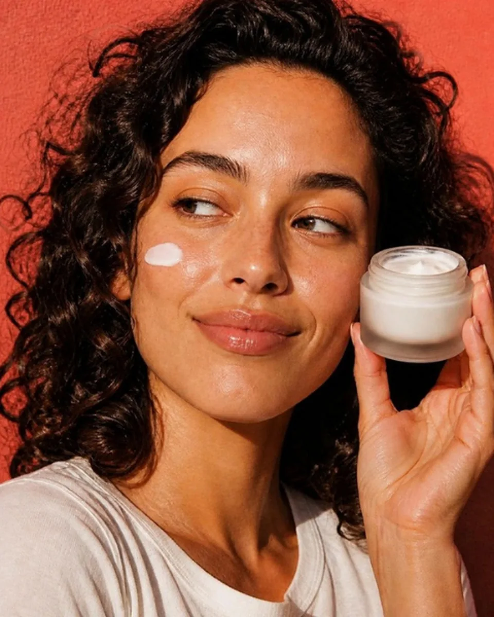 Woman with a dab of moisturizer on her cheek holding a jar of cream beside her face