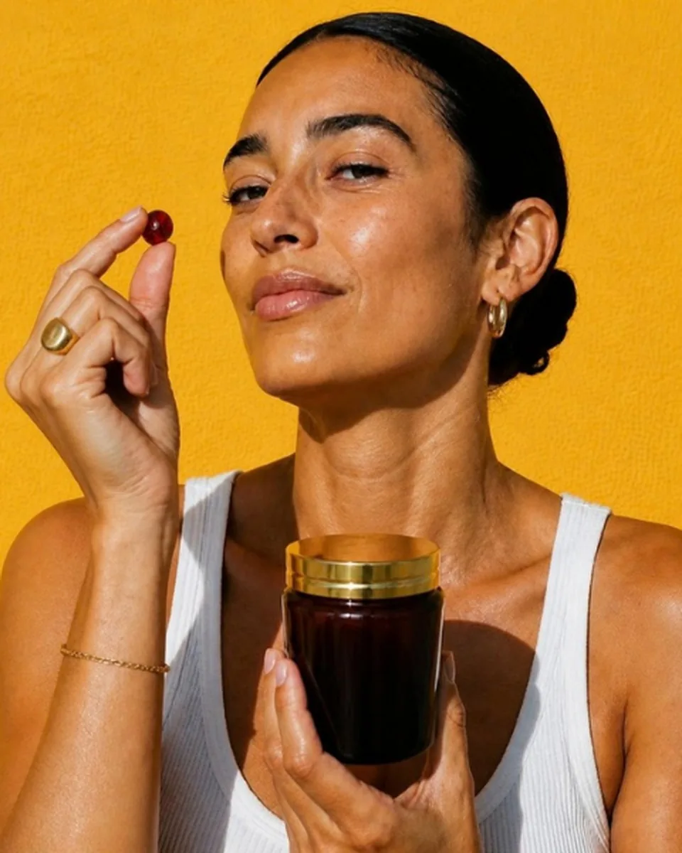 A woman with slicked back hair and gold jewelry holding up a red gummy, with an amber jar with a gold lid in her other hand, on golden yellow