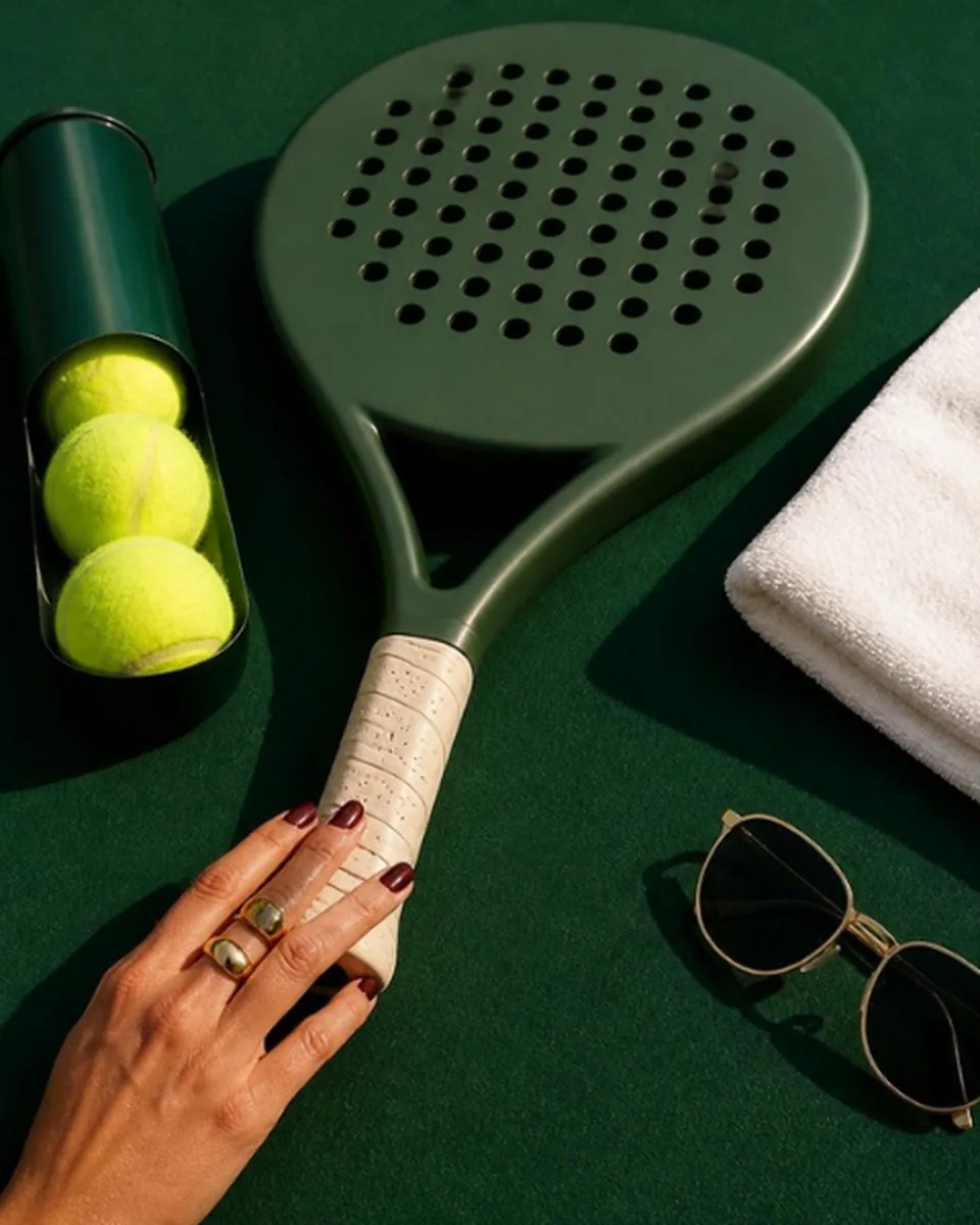 A green padel racket laid flat beside a tube of balls, a white towel and sunglasses, with a hand on the grip